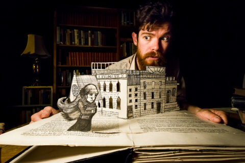 Ralph peers across a book with a building and person cut out in moody lighting
