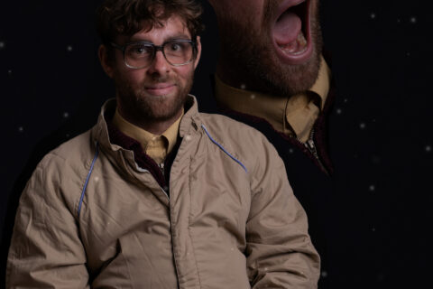 A man is sitting down. He is wearing a beige jacket and reading glasses. The background is black